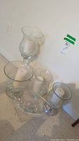 Five glass candle holders placed on carpeted floor, each containing white pillar candles and some with decorative clear glass beads inside. Variations in height and shape from 10 to 22 inches.