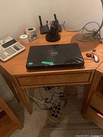 Corner wooden desk with Dell laptop, calculator, pen holder, and mouse on top. Visible drawer and power strip under desk. Chair partially visible.