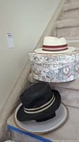 White fedora-style men's summer hat with red band placed on floral patterned hat box on stairs alongside black woven ladies summer hat.