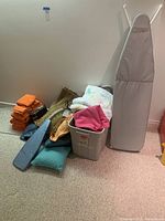 Photo of linens, orange towels, blankets, pillows, a full-sized ironing board and sleeve ironing board, and a storage tote.