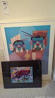 Photo showing two framed prints leaning against a wall. The larger print features a geometric abstract image of two stylized figures in pastel colors. The smaller print is colorful, depicting a house and text, framed in black.