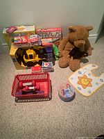Photo of various toys and items including a yellow toy truck, brown stuffed bear, baby bib, collector tin, card decks, marble run, and StrawConnect box.