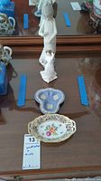 View of two trinket dishes on wooden table along with two porcelain women figurines, one standing and one kneeling.