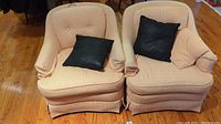 Two matching upholstered tub chairs with armrests, button tufted backs and skirted bases. Each chair holds one black throw pillow and one matching fabric pillow.