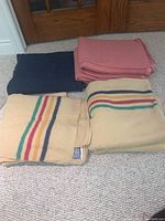 Five wool blankets: two beige with multi-colored stripes, one pink, one dark navy, and one dark blanket in plastic bag on carpet