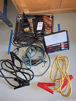 Overview photo showing battery charger, jumper cables, orange ratcheting cargo straps in drip pan, assorted bungees, and wheel chocks