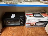 Black & Decker TO1313B toaster oven alongside original packaging box on wooden floor with shelf above containing glassware.