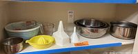 Shelf view showing vintage Pyrex blue striped mixing bowl, Pyrex measuring cups, stainless steel mixing bowls, plastic funnels, and a yellow citrus juicer.