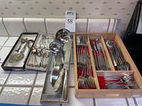 Wide view of flatware and serving pieces arranged in trays and wooden box showing various spoons, forks, and knives with ornate serving spoons in metal trays.