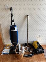 Oreck Magnesium upright vacuum cleaner standing next to a basket with brushes, extra vacuum bags, and vacuum accessories including a metal telescoping wand and hose.