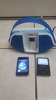 Three iPods displayed with blue and white iLive docking speaker on tile floor