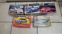 Photo showing all five model car kit boxes together on tiled floor. Boxes include Cadillac Escalade, Dodge Charger, Camaro SS, Thunderbird Turbo Coupe, and Ford Thunderbird.