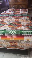 Bed covered by the Pendleton blanket with two matching throw pillows placed near the head of the bed. Pattern includes southwestern geometric shapes in warm earthy colors with green highlights.