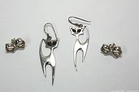 Two pairs of small sterling silver earrings on white background: one pair of stylized cat dangle earrings and one pair of bow-shaped stud earrings.