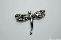 Front view of a silver-tone dragonfly brooch with multicolored inset stones on wings and clear stone on the head, laying on a white background.
