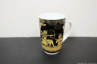 Tall ceramic mug shown from front angle, displaying gold Egyptian figures and hieroglyphs on black background.