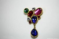 Top view of brooch showing multicolor rhinestones set in gold tone metal, featuring green, red, pink, clear, and blue stones, with a blue teardrop stone hanging.