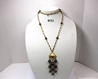 Full view of MYKA necklace displayed on a white bust, showing green beads on gold-tone chain with square pendant and bead strands.