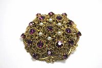 Close up image of vintage gold tone brooch with purple rhinestones and faux pearls showing detailed filigree metalwork and arrangement of stones.