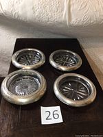 Four round sterling silver-rimmed coasters with clear glass inserts shown from above on a wooden surface, one coaster larger than the other three.