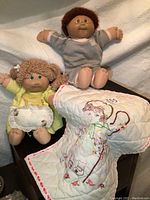 Two Cabbage Patch dolls with quilted embroidered blanket and corner of wooden playpen visible.