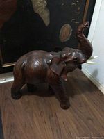 Front side view of large brown leather elephant with raised trunk and visible tusks on wood floor.
