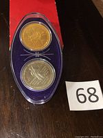 Two commemorative coins in sealed oval plastic case with purple velvet backing placed on a dark surface. A number 68 card is next to the case.