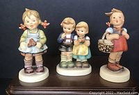 Front view of three Hummel figurines: Little Shopper, Flower Girl, We Congratulate, showing painted details and poses.