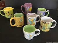 Seven Disney mugs with Tinker Bell designs in various colors and styles, arranged on flat surface