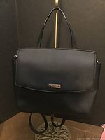 Front view of black Kate Spade handbag showing flap with metal logo plate and permanent top handle.