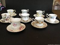 Full lot of 8 tea cups and matching saucers arranged on black surface showing various floral and decorative patterns, all in very good condition with no chips or cracks.