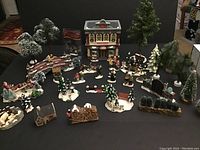 Overview of Christmas village accessories featuring buildings, figurines, trees, and scenic pieces arranged on black surface.