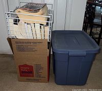 Cardboard box holding multiple books and a white wire crate on top filled with books, alongside a blue plastic tote next to the box.