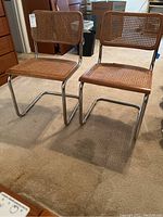 Two rattan and metal chairs side by side on carpeted floor showing overall condition and structure.