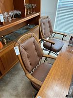 Two wooden dining chairs with brown leather cushions and wooden armrests, set beside a wooden dining table and cabinet with glassware.