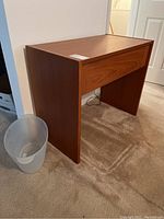 Front side view of the wooden desk on beige carpet with a plastic trash bin next to it.