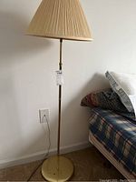 Full view of tall brass floor lamp with cream pleated fabric shade standing next to a bed and wall outlet.