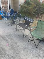 Five different folding chairs arranged in a row outside on concrete with some greenery in background. Includes Coleman chair with black fabric and folding side table, metal and vinyl folding chair, and green fabric folding chair among others.