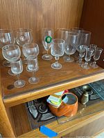 Vintage and clear drinking glasses on wooden shelf, including small liqueur glasses, wine glasses, brandy glasses, some with vintage shapes
