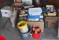 Boxes and containers filled with buckets, cords, tapes, cleaning sponges, and spray bottles arranged on floor