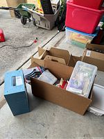 Overall view of cardboard boxes with assorted home repair items including brushes and candles, placed near storage bins in a garage setting.