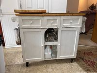 Front view of white wood kitchen island on casters showing three drawers, two cabinets, towel bar, and cutting boards on top.