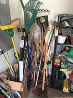 Photo showing assortment of various hand and yard tools clustered inside a green triangular rack, including metal shovels, rakes, hoes, and axes with wooden handles.