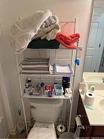 Front view of white wire metal shelving unit positioned over toilet, with various items on shelves for scale. Shelves are empty for sale.