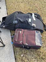 Maroon carry-on suitcase and black golf bag lying on grass. Front and side zippered compartments of suitcase visible.