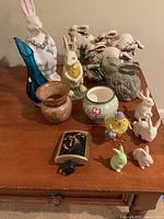 Photo showing various ceramic bunnies, a blue glass vase, two ceramic vases (brown and floral), a yellow chick figurine, and a small nightlight on a wooden surface.