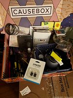 Full view of the Causebox subscription box filled with various items including an Anker USB adapter, small tins, a glass tumbler, and assorted small items.