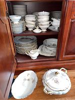 View of open cabinet showing stacks of dinner plates, small plates, bowls, cups, saucers, gravy boat, and creamer inside cabinet, plus covered soup tureen and serving platter on table surface in front.