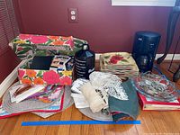 Wide view of all lot items including Krups coffee pot, stack of floral square plates, assorted kitchen linens, placemats, oversized water bottle, note chips, and decorative items on floor against wall