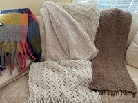 Four decorative throws displayed on a couch: colorful plaid, beige sherpa, brown chunky knit, and cream open knit